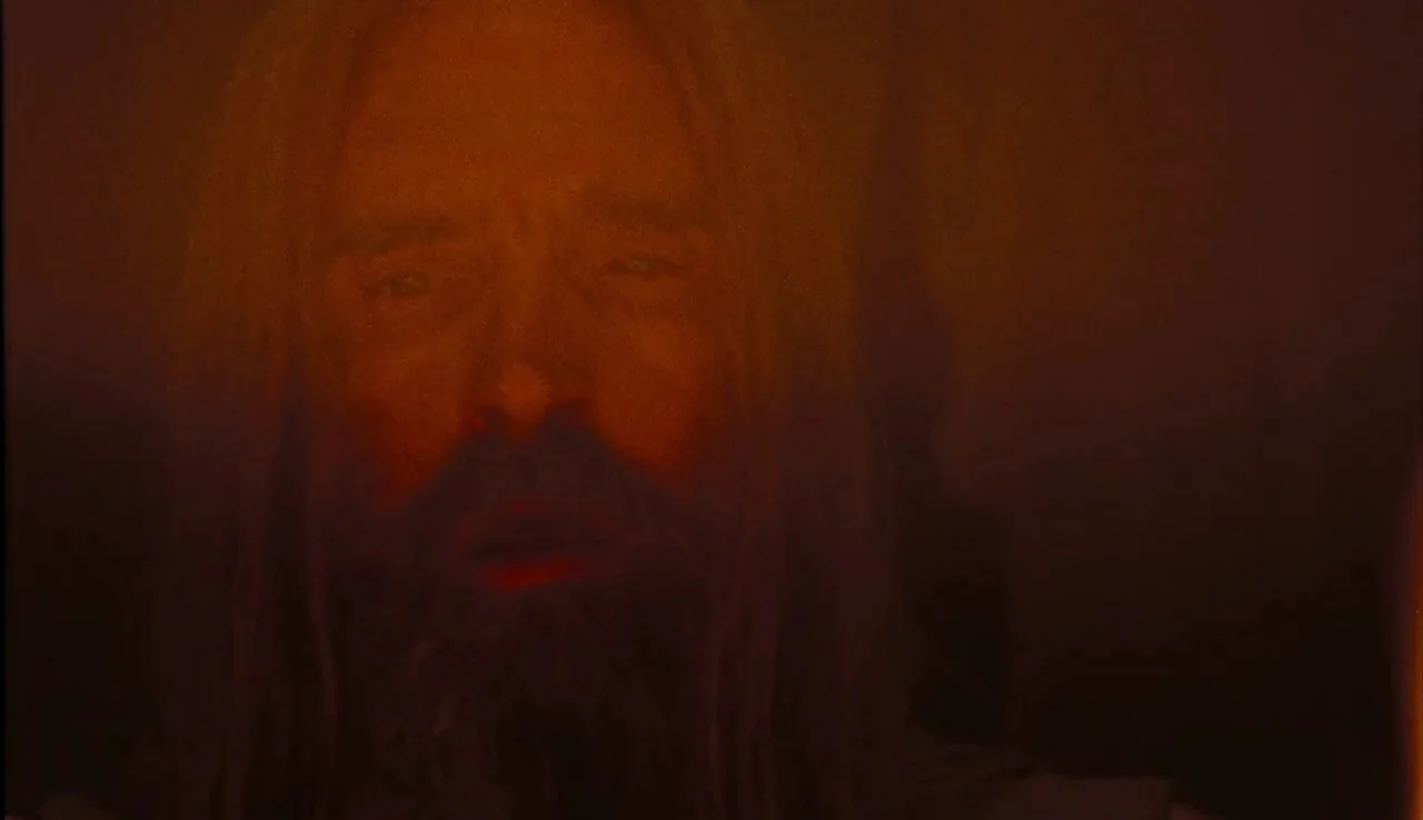 The Devil’s Rejects (2005) movie still: closeup — A man's face emerges from a dense, dark, amber-tinted environment. The lighting emphasizes his…