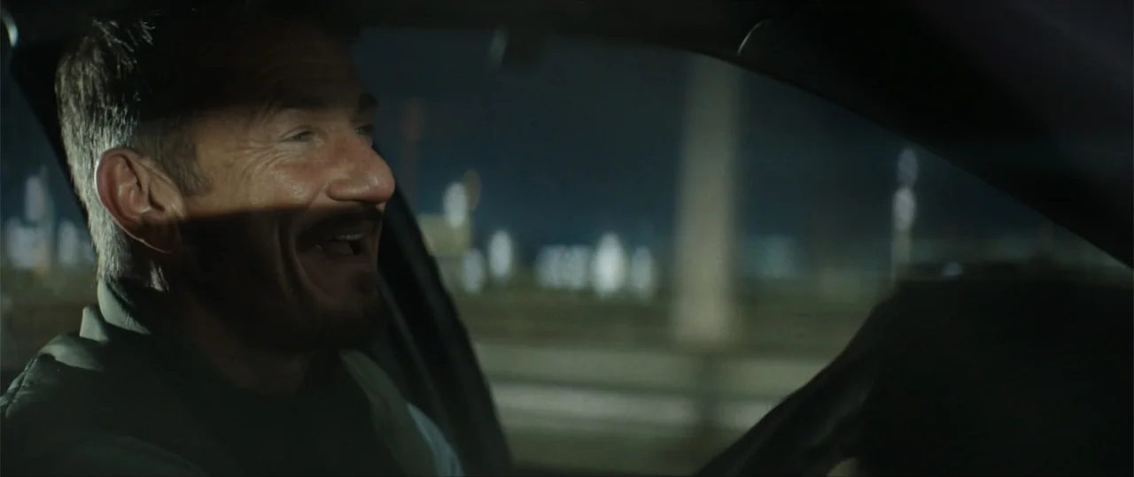 Daddio (2024) movie still: closeup — A man drives a car at night with city lights blurred in the background. He looks to his right while smiling…