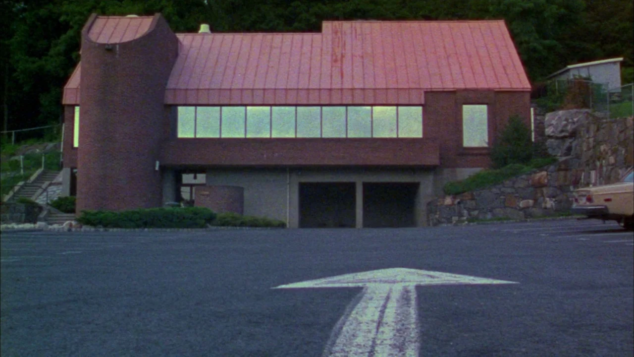 Poison movie still: establishing — A brick commercial building with a metal roof overlooks a paved asphalt parking lot. A large white directional…