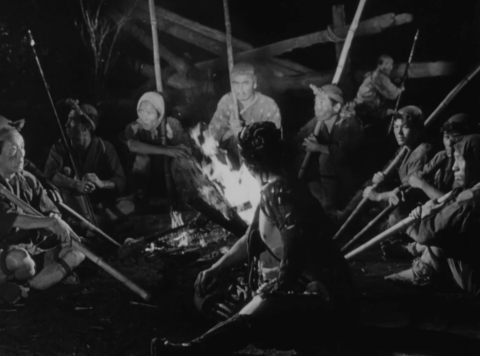 Seven Samurai (1954) movie still: medium — A group of men sit around a fire in a dark, wooded outdoor clearing. They hold long wooden spears while…