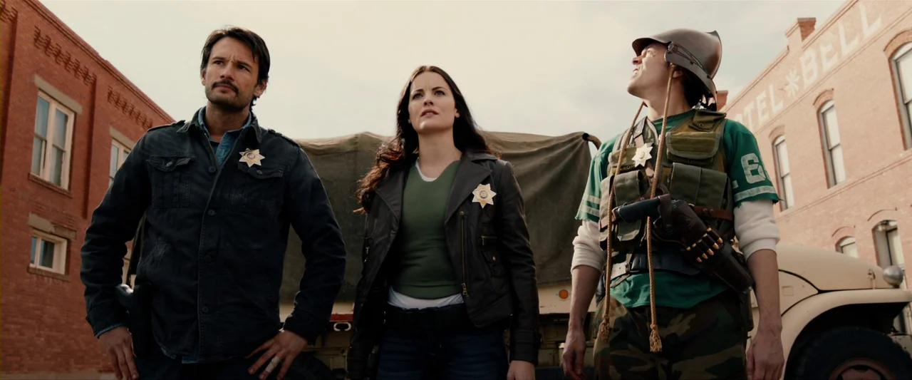 The Last Stand (2013) movie still: fullbody — A group stands on a town street between two red brick buildings. A man and woman in badges and a man…