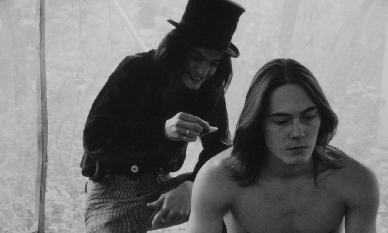 Multiple Maniacs (1970) movie still: medium — A person wearing a black top hat stands inside a light-colored canvas tent. They are tending to the…