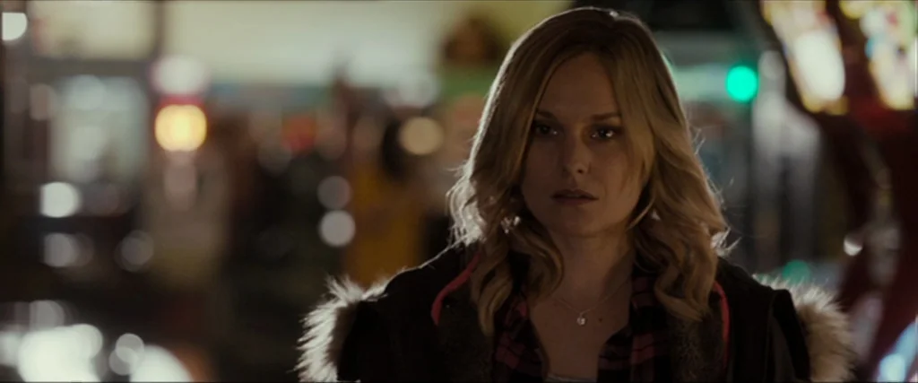 Clown (2014) movie still: medium — A woman stands before a blurred background of colorful night carnival lights. She gazes ahead with a neutral…
