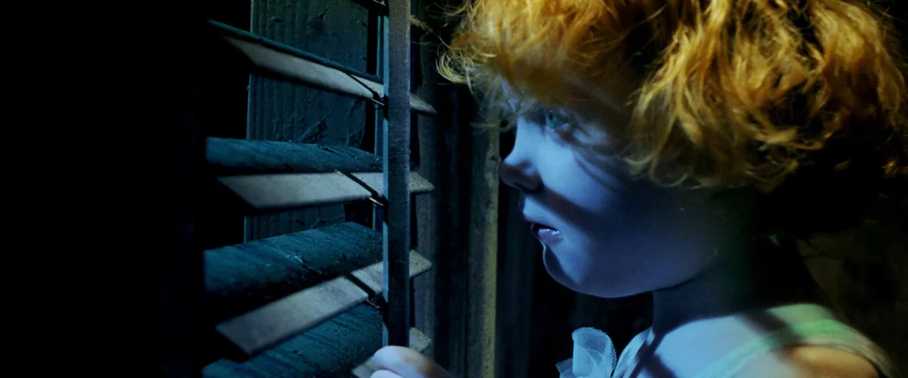 Moon Garden (2022) movie still: closeup — A dark room features a young girl peering through wooden window blinds. She presses her face close to the…