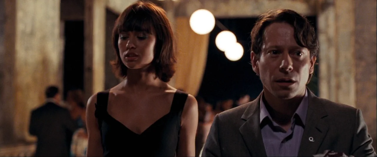 Quantum of Solace (2008) movie still: medium — A spacious hall with tall columns and distant light fixtures hosts a gathering. A man in a suit and a…