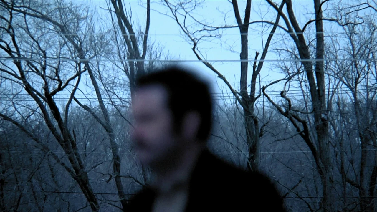 A Horrible Way To Die (2010) movie still: medium — A man moves past a cluster of bare trees during the late afternoon. Utility lines stretch…