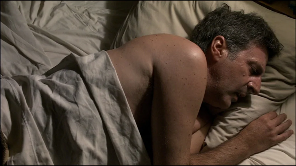 Caché (Hidden) (2005) movie still: medium — A man sleeps in a plain bed with cream-colored sheets and a pillow. He is covered from the waist down…