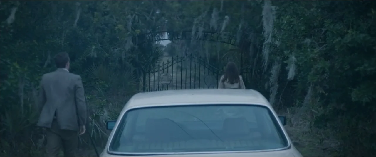 Offseason (2022) movie still: wide — A man and a woman walk toward an old iron gate between dense trees. The metal gate stands at the entrance of a…