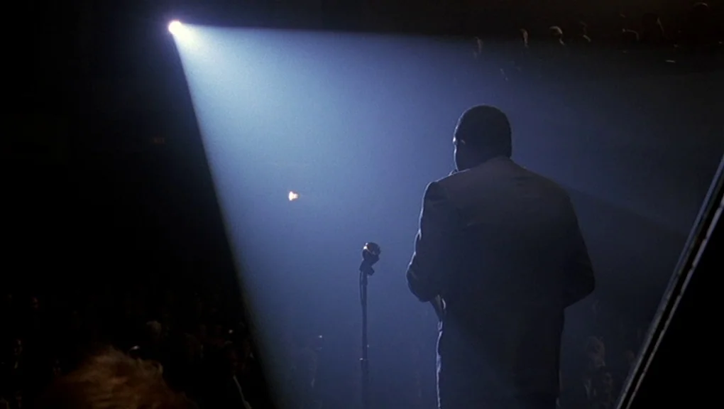 Bird (1988) movie still: medium — A man in a suit stands on a dark podium in an auditorium. He holds a microphone while a single harsh spotlight…