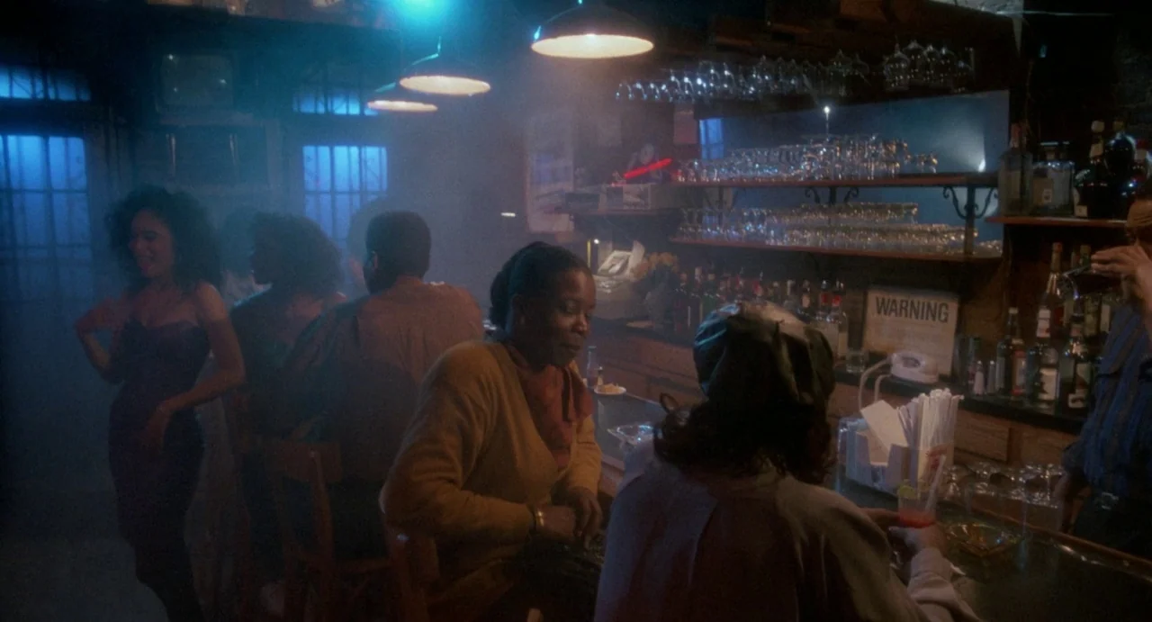 Def By Temptation (1990) movie still: medium — A dimly lit drinking establishment features a long wooden counter and rows of glassware. Several…