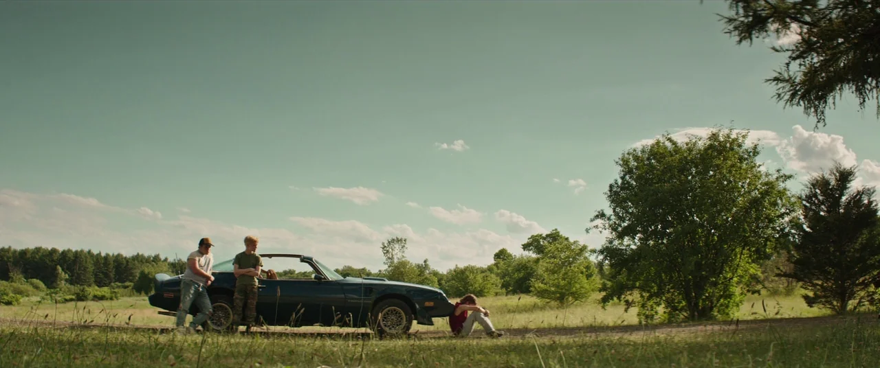 It (2017) movie still: wide — An open rural field features a dark blue sports car parked on a dirt road. Three men gather around the vehicle under a…