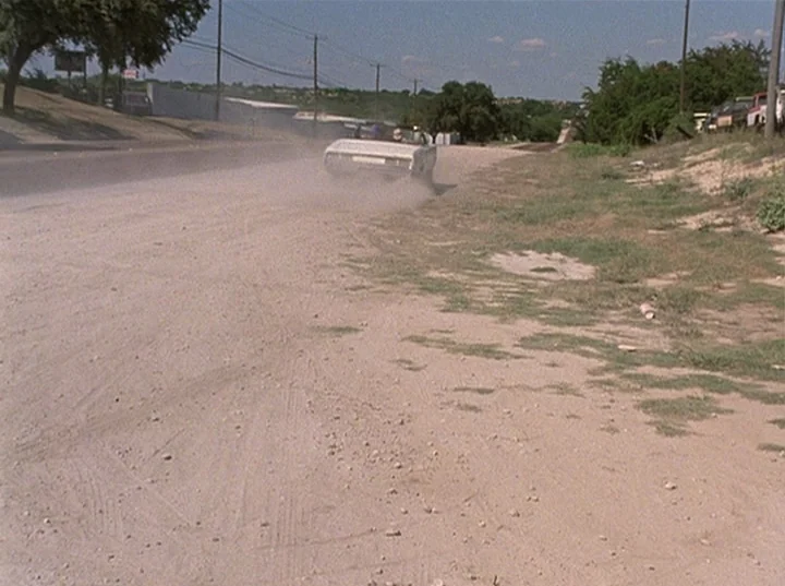 Slacker (1991) movie still: wide — A dirt shoulder borders a paved road in a sparsely populated rural area. A light-colored convertible car drives…