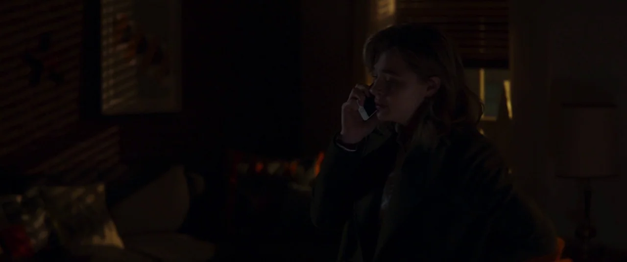 Greta movie still: medium — A dimly lit room with closed window blinds casting shadow stripes on the wall. A woman stands in the dark space talking…