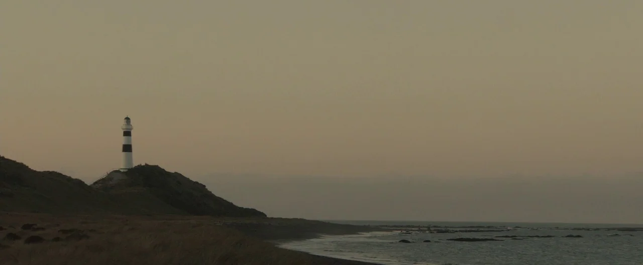 The Light Between Oceans (2016) movie still: wide — A tall white lighthouse with black bands stands on a dark, grassy coastal hill. The ocean meets…