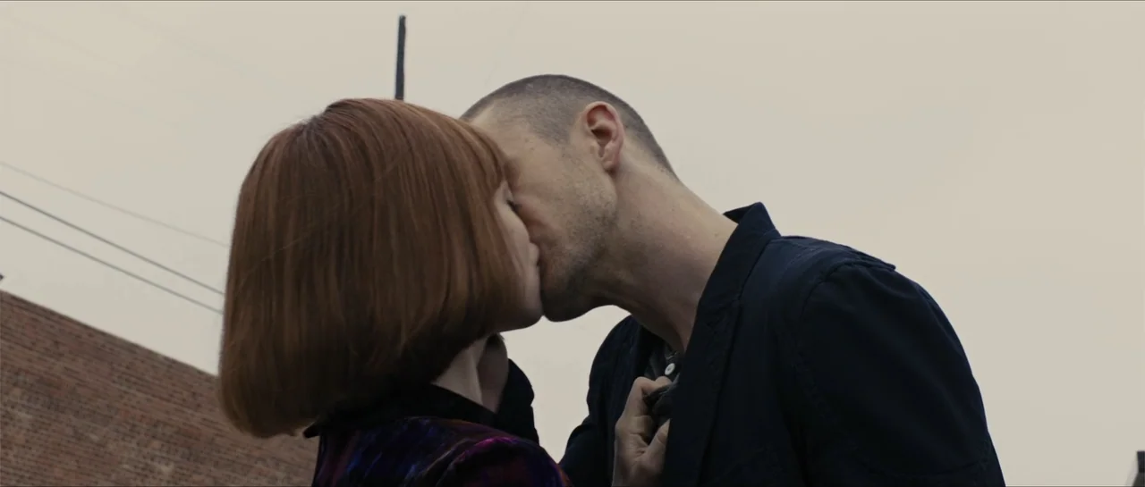 In Time (2011) movie still: medium — An exterior rooftop with a flat gray sky and a brick wall visible. A man and a woman stand close together and…