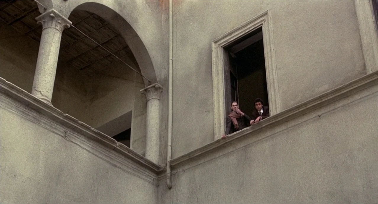 Amarcord (1973) movie still: wide — A stone courtyard features high walls and arched columns in an old building. Two men stand at an upper-level…