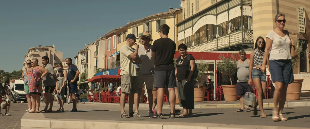 The Only One (2021) movie still: fullbody — A sunny Mediterranean waterfront promenade features pastel buildings and red outdoor cafe furniture.…