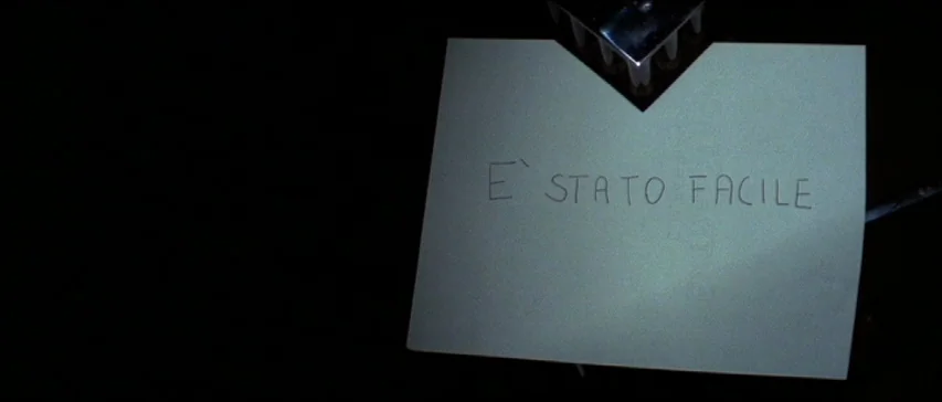 Four Flies On Grey Velvet (1971) movie still: closeup — A piece of paper with hand-written Italian text lies on a dark surface. The message reads, E…