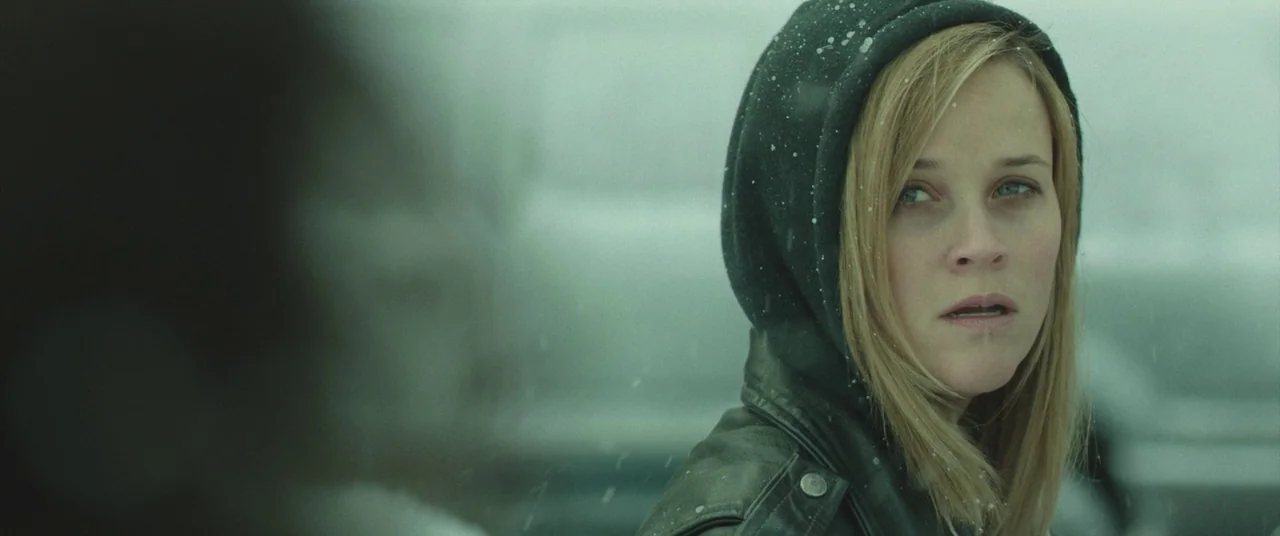 Wild (2014) movie still: closeup — A snowy outdoor setting with soft diffused light and blurred gray background elements. A woman in a black hooded…