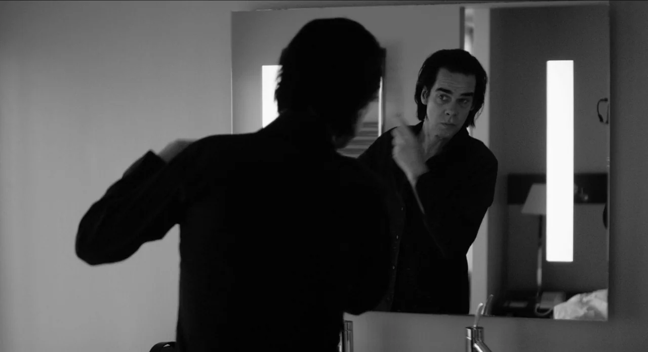 One More Time With Feeling movie still: medium — A dimly lit room contains a large rectangular wall mirror with integrated vertical lighting. A man…