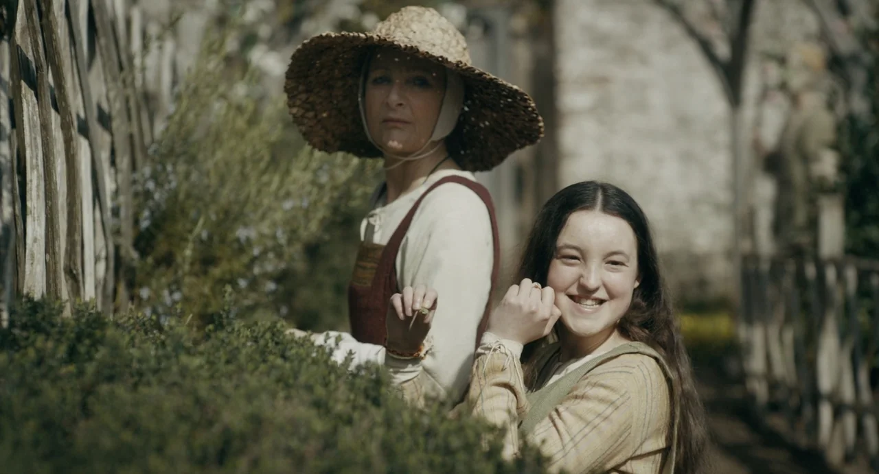 Catherine Called Birdy (2022) movie still: medium — A rural outdoor space featuring a rustic wooden fence and lush greenery. An adolescent girl…