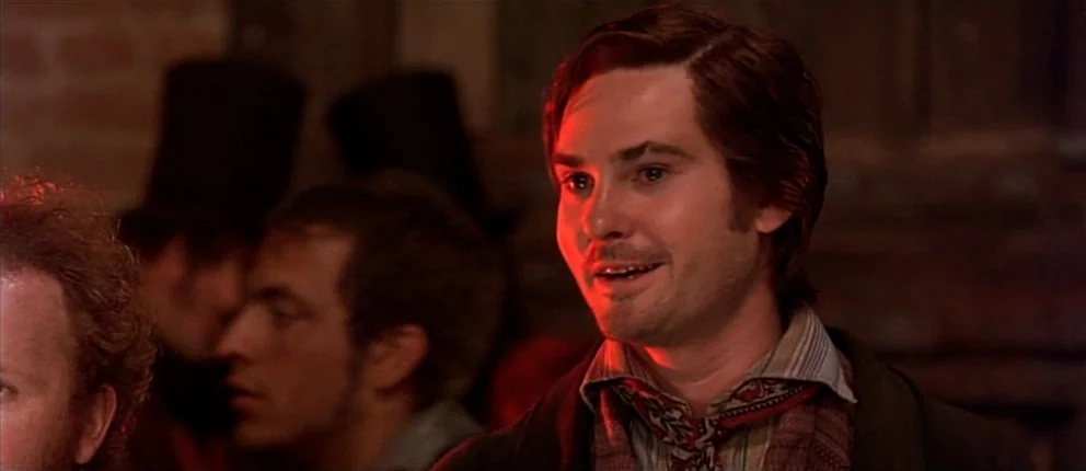 Gangs of New York (2002) movie still: closeup — A dimly lit room with blurred figures in the background near a brick wall. A man in period attire…