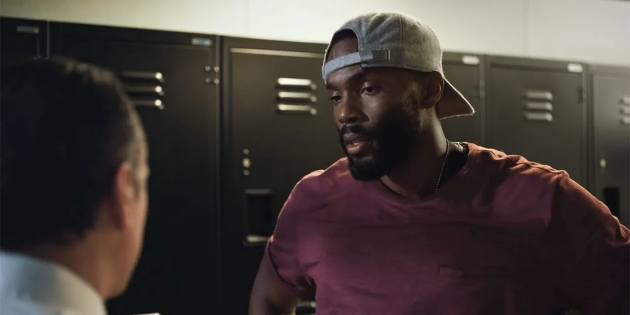 Equal Standard (2021) movie still: medium — A locker room with a row of black metal lockers lining the back wall. A man in a maroon t-shirt stands…