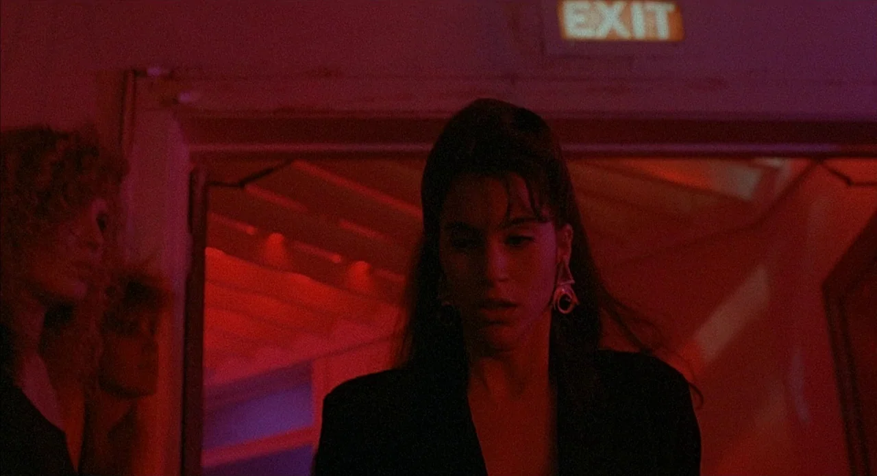 Less Than Zero (1987) movie still: medium — A dimly lit room illuminated by intense red neon light from an exit sign. A young woman with dark hair…