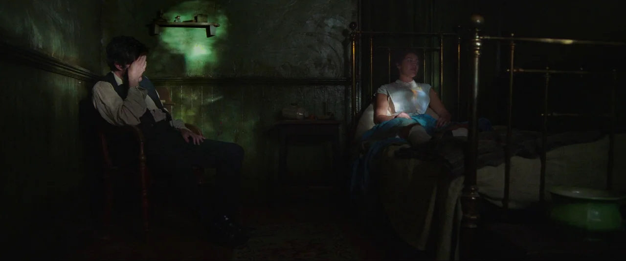 The Wonder (2022) movie still: wide — A dimly lit room with textured dark walls and a brass bed frame. A man sits slumped in a chair while a woman…