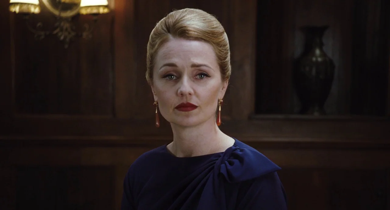 Sleeping Beauty (2011) movie still: closeup — A woman stands centered against dark wood paneling with wall lamps visible. The woman looks toward the…