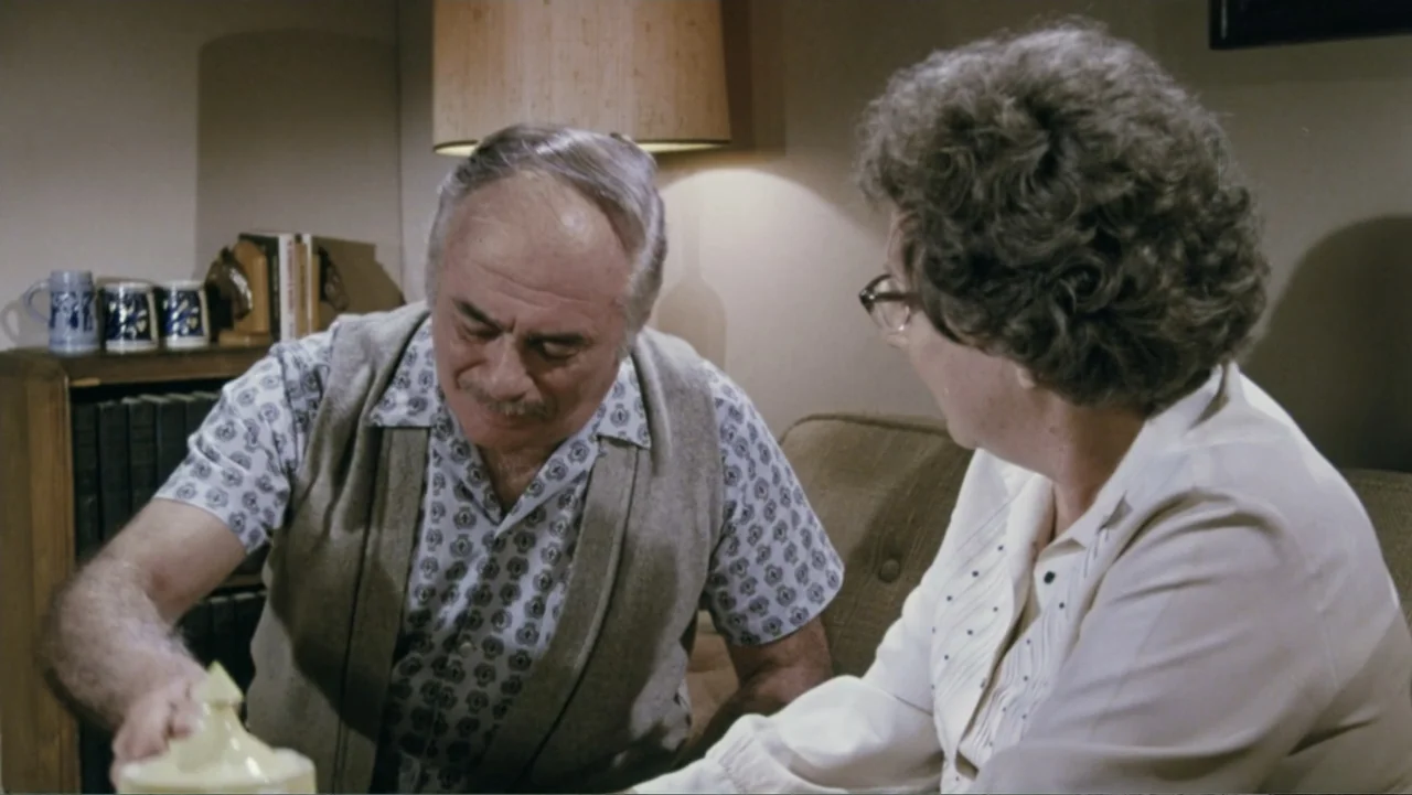 Aunt Mary (1979) movie still: medium — A dimly lit room contains a shelf with mugs and books in the background. An older couple sits together as the…