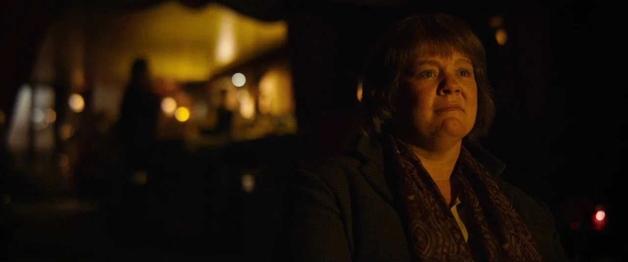 Can You Ever Forgive Me? (2018) movie still: medium — A dark dimly lit pub with blurred figures in the background. A middle-aged woman sits at a…
