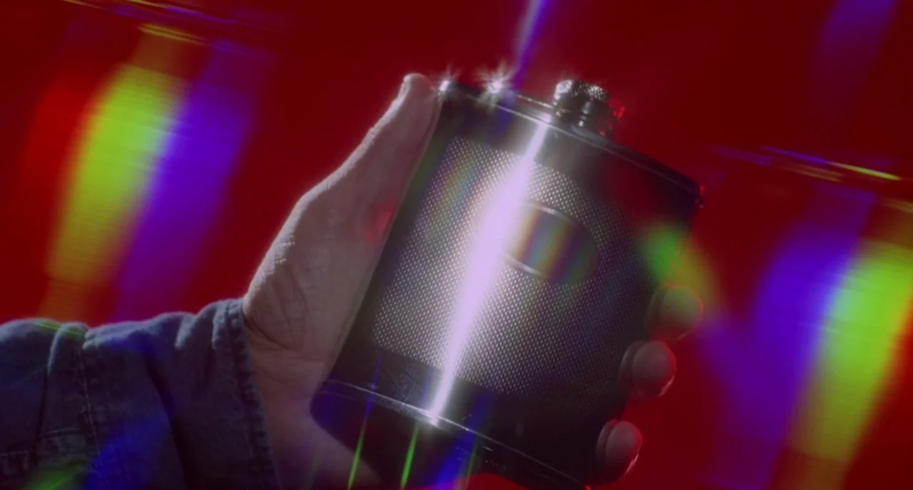 The Love Witch (2016) movie still: closeup — A close-up shot of a hand holding a textured black metal flask. A dark red background is overlayed with…
