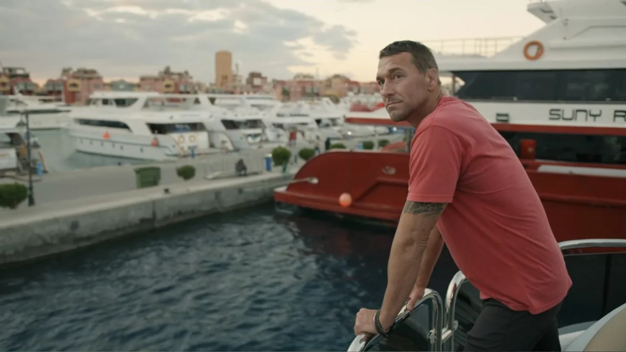 Serial Killer: Red Sea Attacks (2023) movie still: medium — A man stands on the deck of a boat moored at a crowded marina. He leans against the…