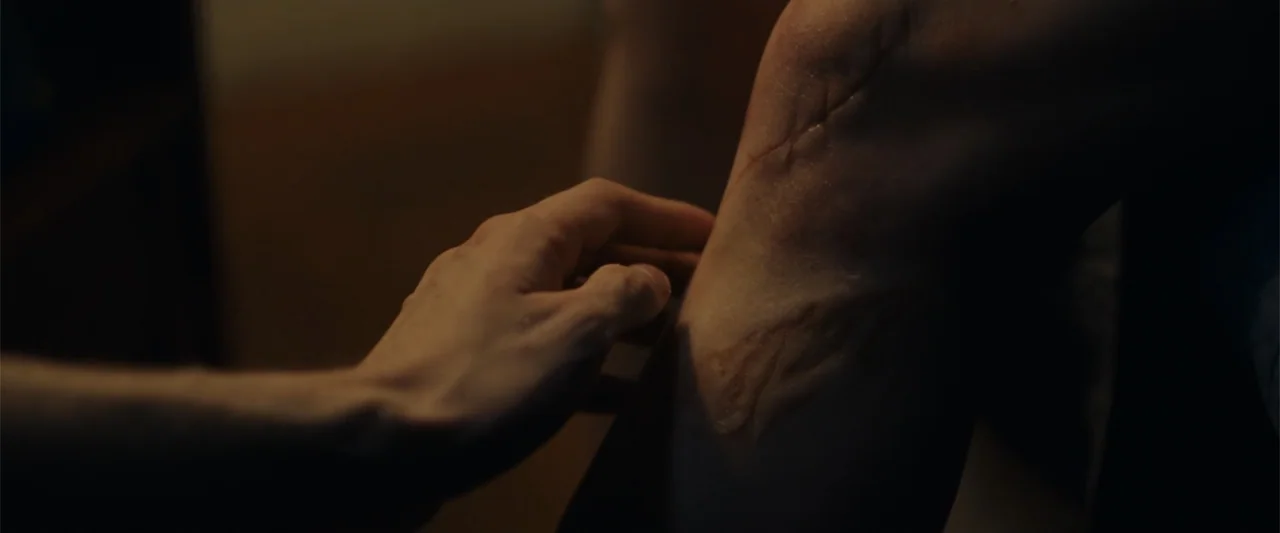 Chemical Hearts (2020) movie still: closeup — A dimly lit interior space focuses on a person's knee and an accompanying hand. A detailed, raised…