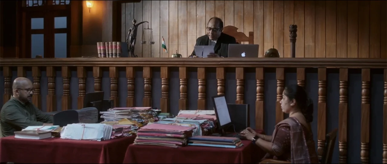 Garudan (2024) movie still: wide — A courtroom features a raised bench with wood paneling and red-clothed desks for clerks. A judge reads a paper…
