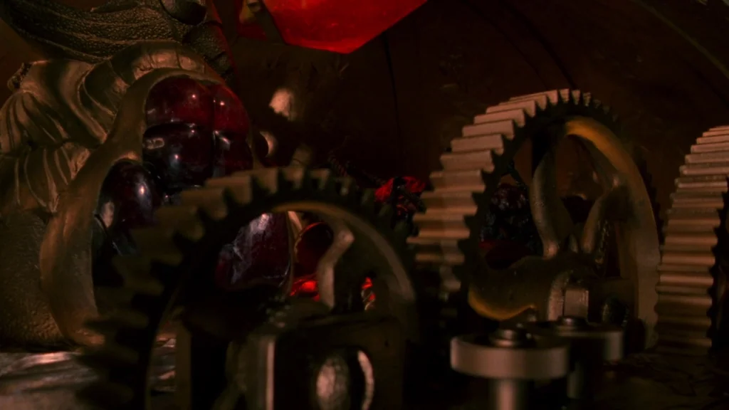 Cronos (1993) movie still: closeup — A dark interior filled with heavy iron gears and intricate mechanical clockwork components. Deep red light…