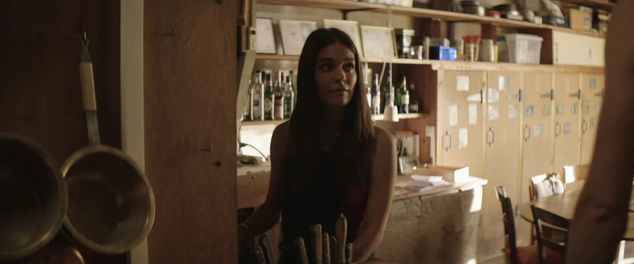 The Only One (2021) movie still: medium — A wooden bar counter cluttered with knife blocks and background shelving holding bottles and awards. A…
