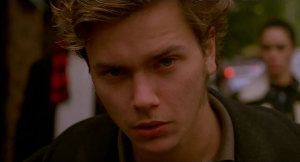 My Own Private Idaho (1991) movie still: closeup — An urban street scene with blurred architectural elements and foliage in the distance. A man in…