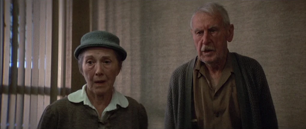 3 Women (1977) movie still: medium — Two elderly people stand in a room with neutral-toned walls and vertical blinds. They face forward with serious…