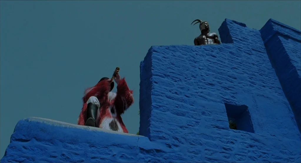 The Fall movie still: wide — A bright blue stone wall structure rises against a clear blue sky. A man in a fur coat looks through a telescope while…