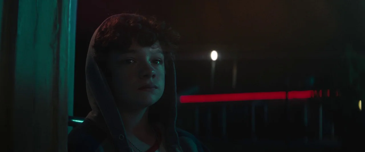 Honey Boy (2019) movie still: closeup — An exterior view of a motel corridor at night illuminated by neon signage. A teenage boy in a hooded…