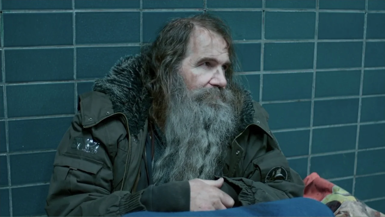 Code of Trust (2019) movie still: medium — A man sits against a wall of dark blue square tiles. He wears a heavy winter jacket and looks toward the…