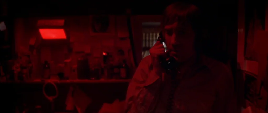 The Omen (1976) movie still: medium — A man stands in a darkroom lit by a red safelight lamp. He holds a black telephone receiver to his ear during…
