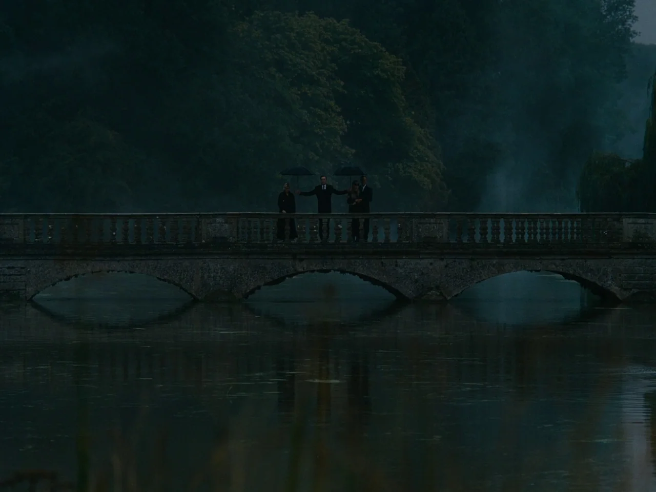 Saltburn (2023) movie still: wide — A three-arched stone bridge crosses a dark, still body of water amid foggy woodland. Three people stand…