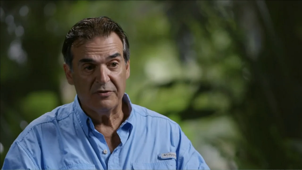 Finding Amelia (2024) movie still: medium — A man stands outdoors in front of a blurry background of green foliage. The man talks to someone…