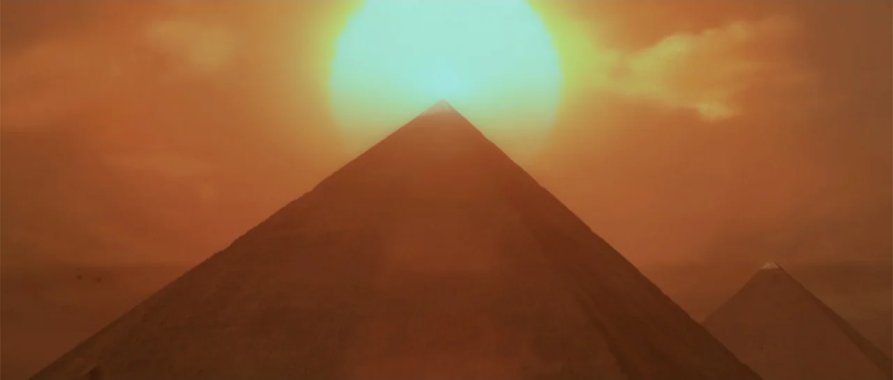 The Mummy (1999) movie still: establishing — Two large stone pyramids rise from a flat desert landscape under a hazy sun. The bright light…