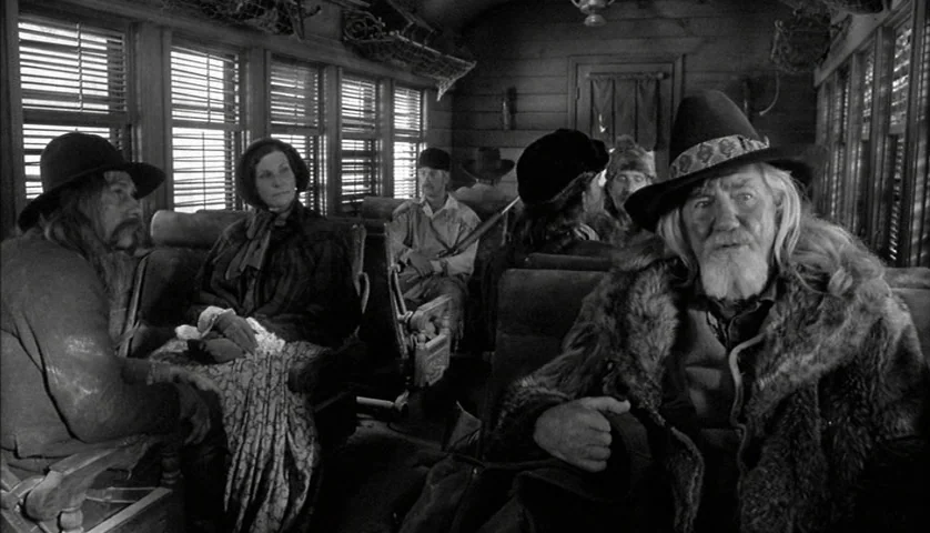 Dead Man (1995) movie still: medium — A vintage wooden train carriage with slatted window blinds and rowed seating. Several passengers in historical…