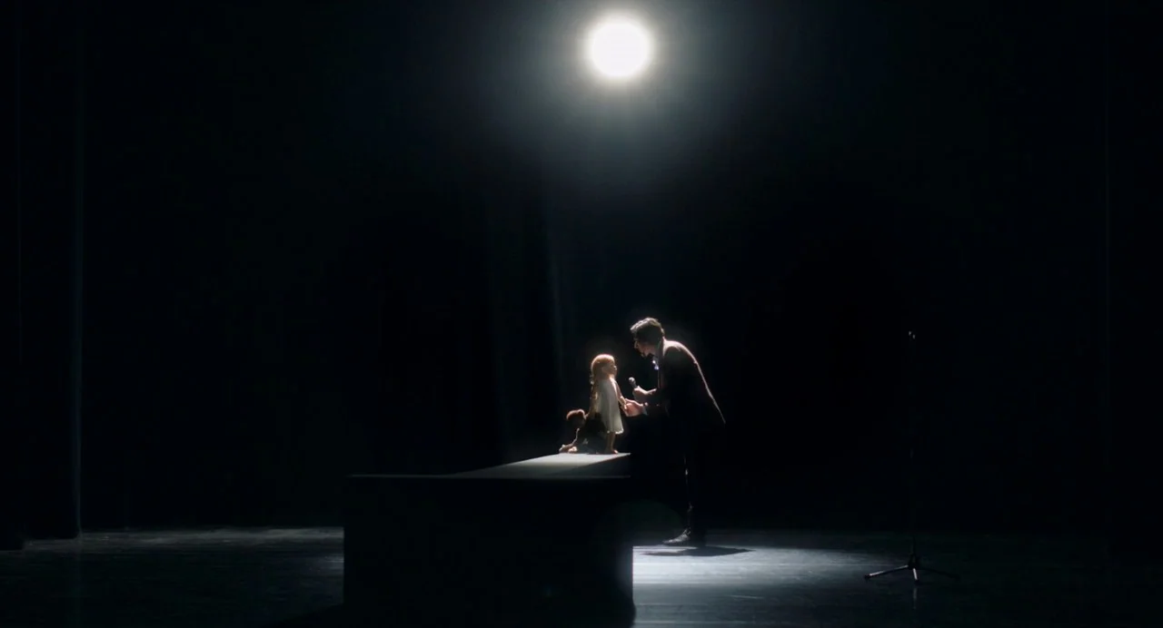Annette (2021) movie still: wide — A dark empty theater stage illuminated by a single high-intensity overhead spotlight. A man in a suit offers a…