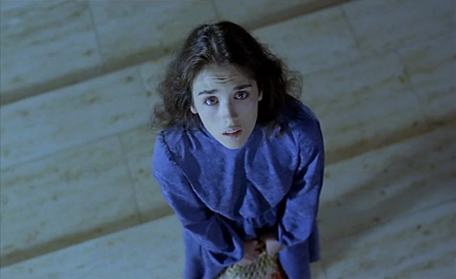 Possession (1981) movie still: medium — A woman stands in a beige stone-tiled stairwell looking directly upward. She wears a blue dress and holds a…